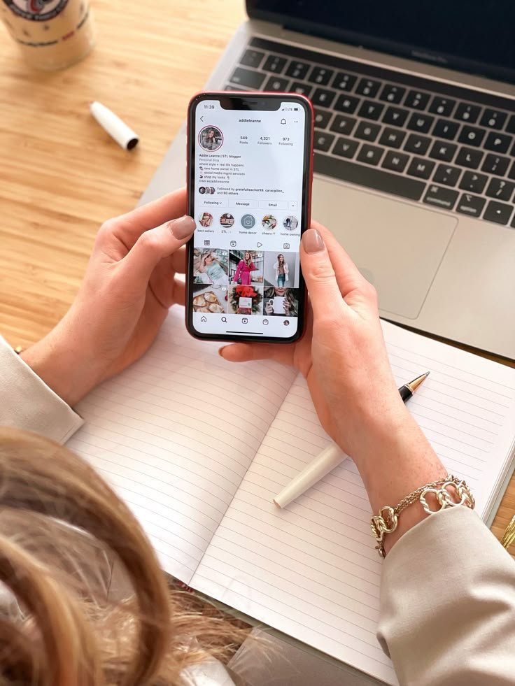 A person holding a smartphone showing an Instagram profile while working on a laptop and notebook — representing creative strategies to make your brand stand out on social media.